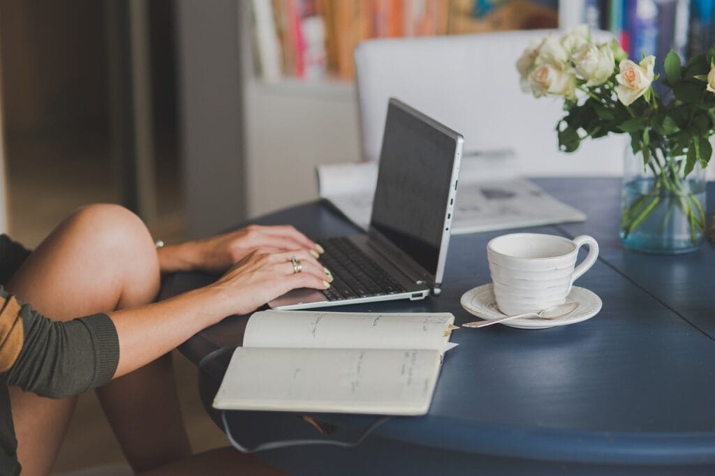 Pessoa escrevendo em notebook com caderno e xícara de café sobre a mesa, representando foco e produtividade em ambiente caseiro.
